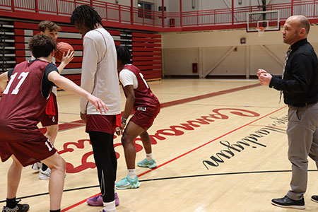 Varsity basketball coach stands nearby as players practice on the court