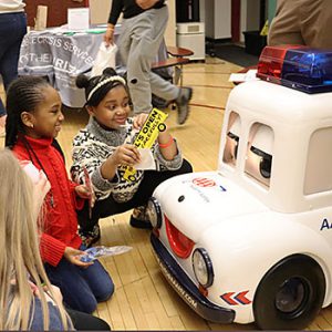 Two students are smiling and laughing as they kneel on the floor next to Otto the Auto robot.