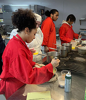 A student wearing a red chef's smock stirs a bowl of ingredients an instructor holding a measuring cup stands nearby  