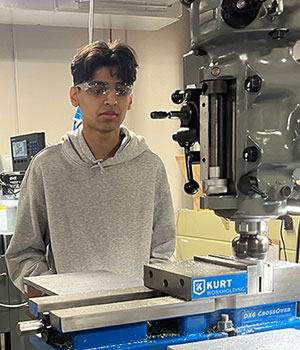 A student wearing protective eye gear feeds a piece of lumber into a machine 