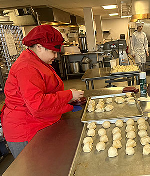 A students wearing a red smock and red chef's hat rolls dough into balls to place on two baking sheets on an adjacent table 