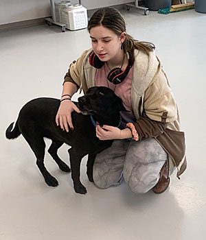 A student kneels on the tile floor and pets a black dog