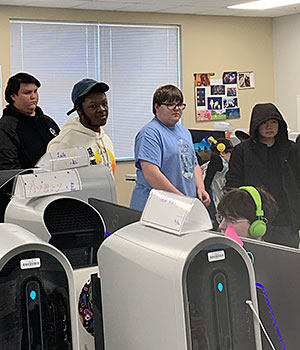 A group of four students observes another student with headphones who is seated at a computer 
