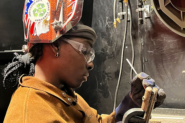 Student wearing protective eye gear, a helmet and thick gloves works with welding tools. 