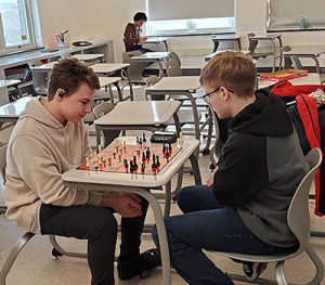 Two students sit on opposite sides of a desk with a chessboard between them.