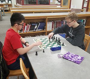 Two students sit on opposite sides of a library table with a chessboard between them. One student moves a chess piece as the other student watches.