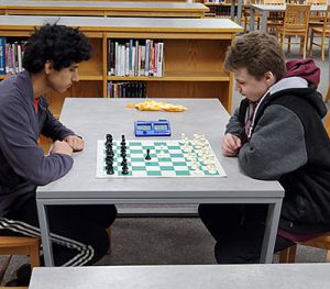 Two students sit on opposite sides of a library table with a chessboard between them.