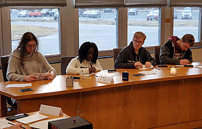 Four members of the Masterminds team sit at a long table with pens in hand, listening and scribbling notes on paper. 