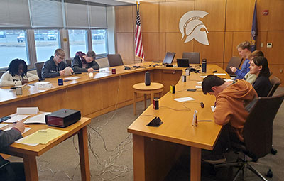 Four members of the Masterminds team sit at a long table opposite another team. Many of the team members are holding pens in hand, listening and some are scribbling notes on paper. 