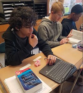 A student sits at a desk with his left hand on the keyboard of a Chromebook. A dictionary and pink baby carriage are on the desk in front of the student. 