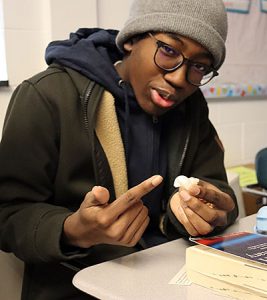 A student wearing a knit cap and glasses with dark frames, points to the snail shell he is holding.