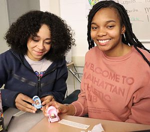 Two students sitting at neighboring desks smile at the camera. One student holds a blue baby carriage, the other holds a pink carriage. Small snail shells peek out from the carriages. 