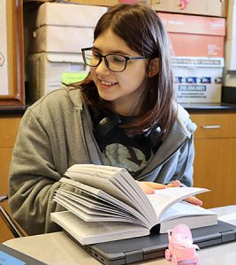 A student with glasses seated at a desk thumbs through a dictionary as a pink baby carriage sits in front of a nearby Chromebook