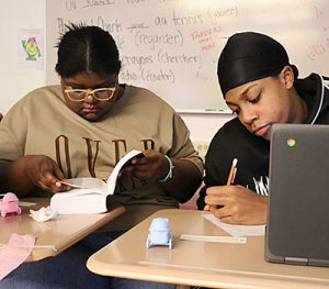 Two students sit at neighboring desks. One students looks down at an open dictionary and a pink baby carriage nearby on the desk. The other students has an open Chromebook and is writing with a pencil on paper. A blue baby carriage is nearby on the desk.  