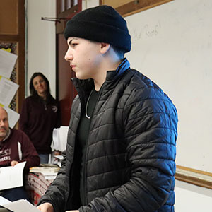 A student dressed in a black jacket and knit cap holds a piece of paper and looks forward. Two educators are standing near the classroom door in the background.  