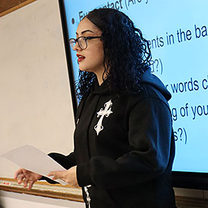 A students wearing glasses and dressed in a black hoodie sweatshirt holds a piece of paper. A whiteboard is on the wall behind the student.