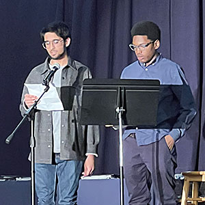 One student reads from a piece of paper into a microphone as another student stands nearby with his hands in his pockets. 