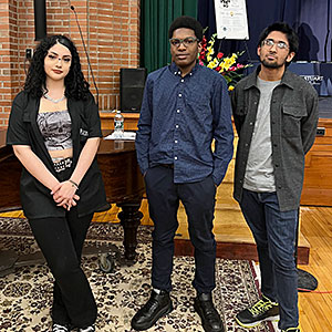 Three high school students stand together with a piano, a stage and flowers in the background. The students are facing the camera.