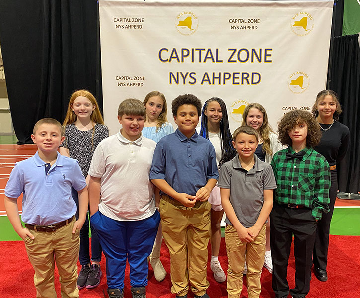Ten elementary students stand in front of Capital Zone Physical Education Leadership award banner