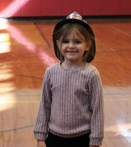 A young student wears a black plastic fireman's hat and smiles