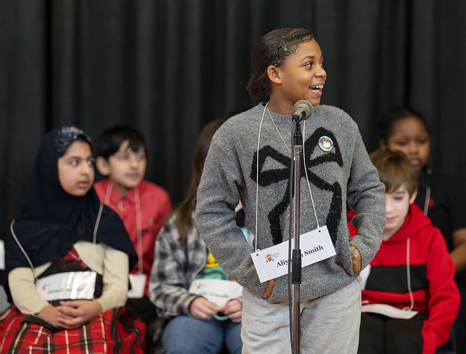 A runner up stands at the microphone and glances toward the pronouncer's podium and smiles