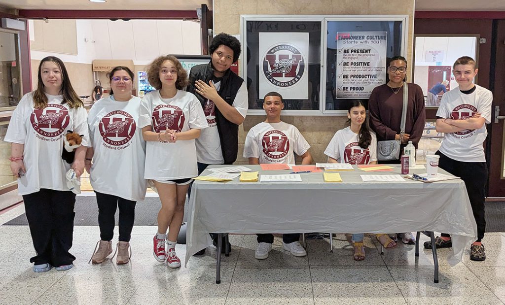 A group of students stand by or sit at a table to welcome Career Fair guests