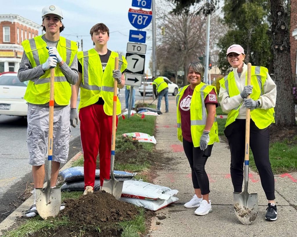 Two character education students stand with two advisors with shovels in hand and bags of top soil at their feet 