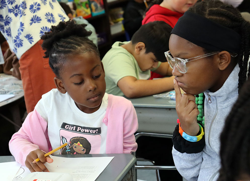 An older student sits at a desk with a younger student as the two talk to each other.
