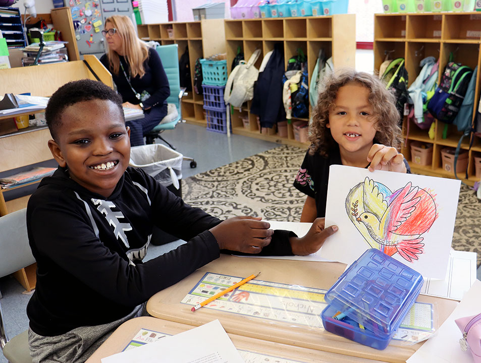 An older students smiles as a younger student holds up a paper peace dove