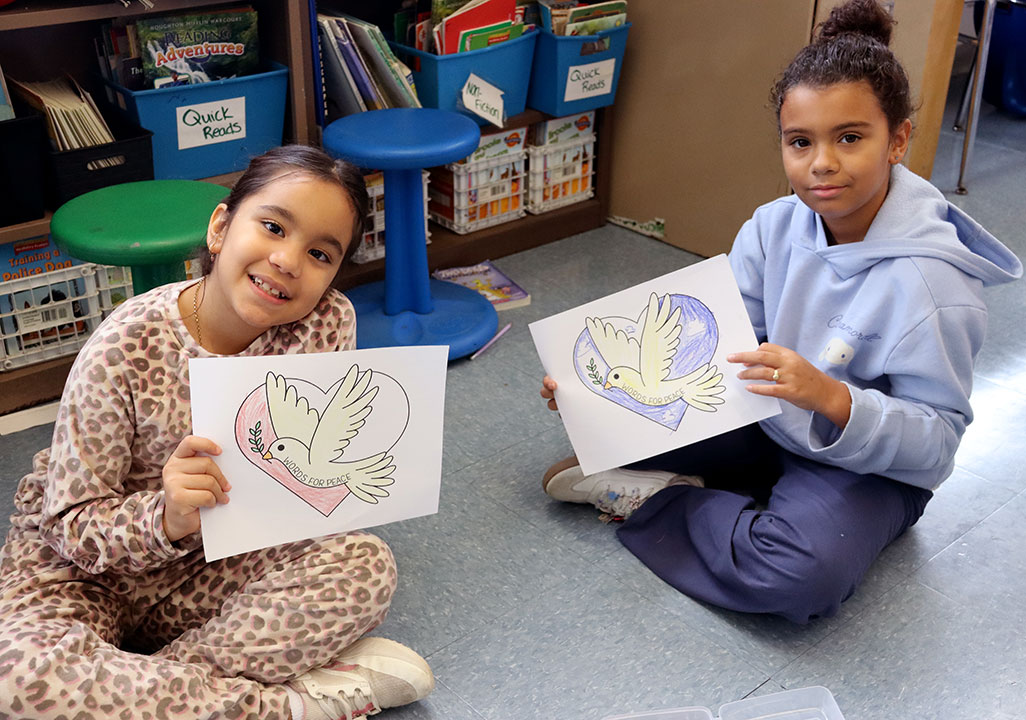Two students seated on the floor hold up paper peace doves.