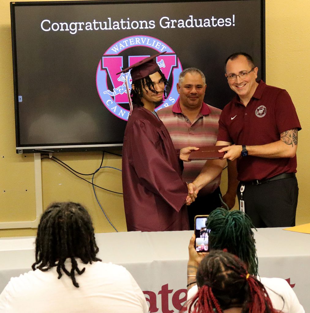 The vice president of the school board looks on as a student shakes the superintendent's hand while accepting their high school diploma.