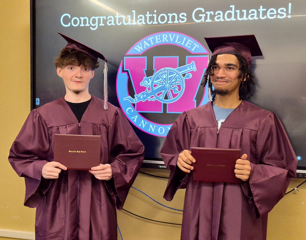 Two students dressed in caps and gowns hold their diplomas and smile