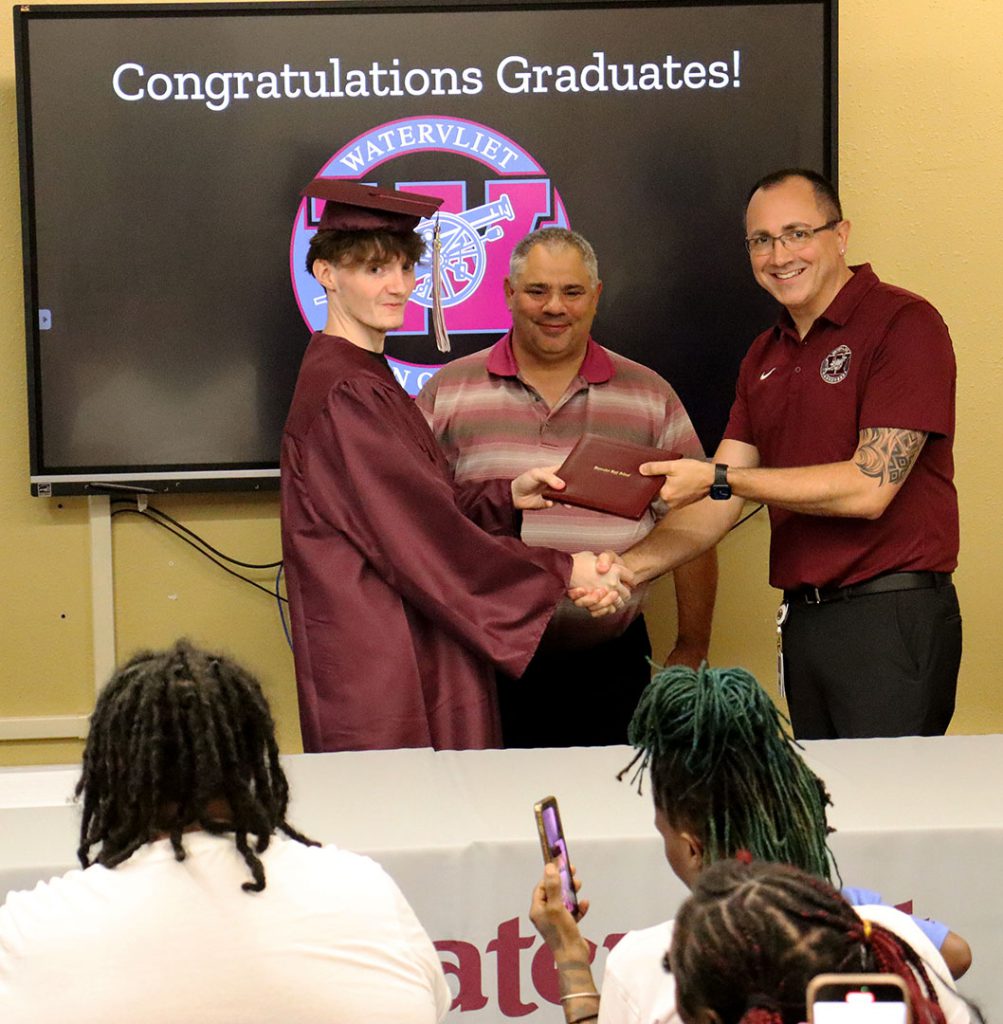 The vice president of the school board looks on as a student shakes the superintendent's hand while accepting their high school diploma.